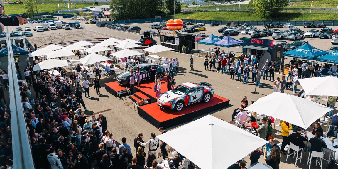 Zwei Porsche Sportwagen auf einem Podest umringt von Porsche Cars & Coffee Gästen.