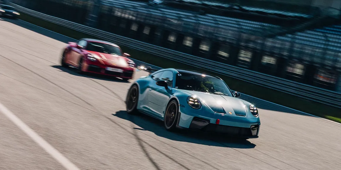 Ein türkiser Porsche im Vordergrund fährt mit einem roten Porsche im Hintergrund auf einer Rennstrecke